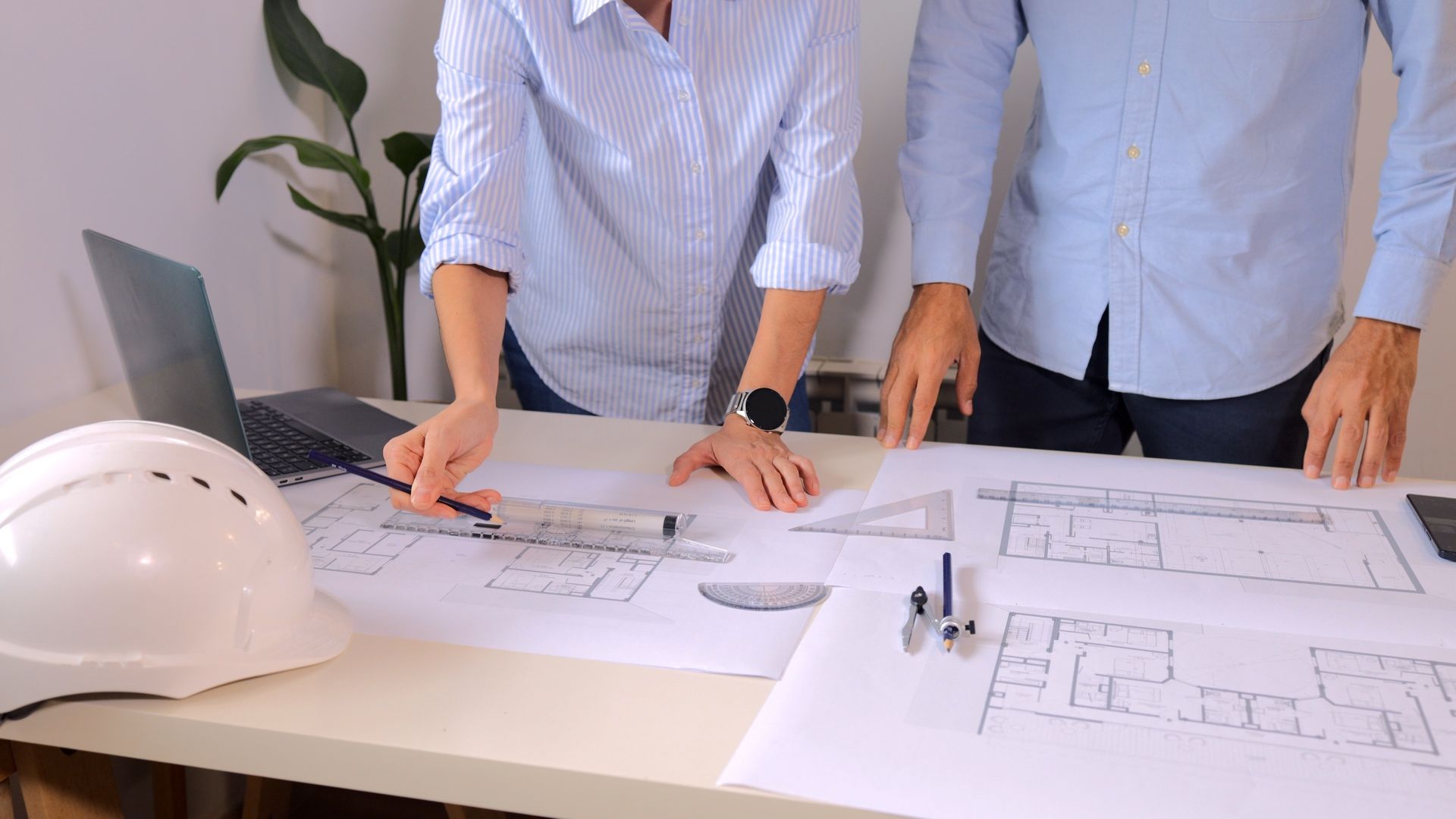 Two people reviewing architectural blueprints on a table with a white hard hat nearby.