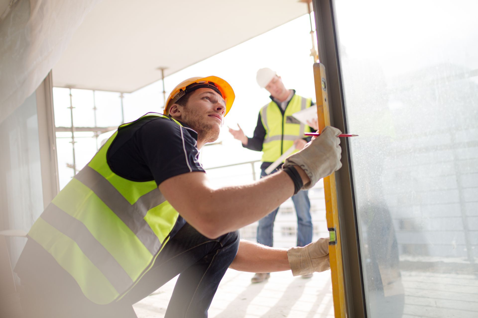Two construction workers in safety vests installing a window panel at a bright indoor site