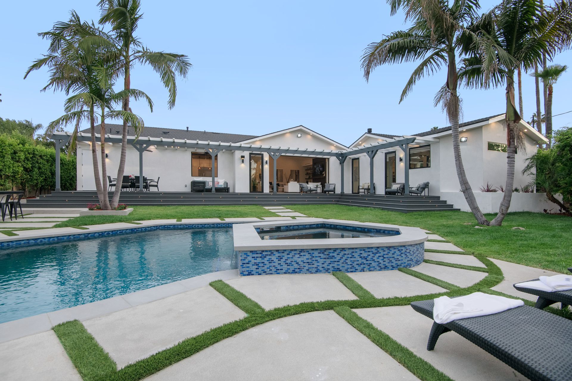 Luxury backyard with a pool, hot tub, palm trees, and a white modern house under a clear sky