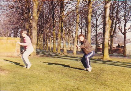 Two men are squatting in a park with trees in the background practicing Tai Chi Chuan