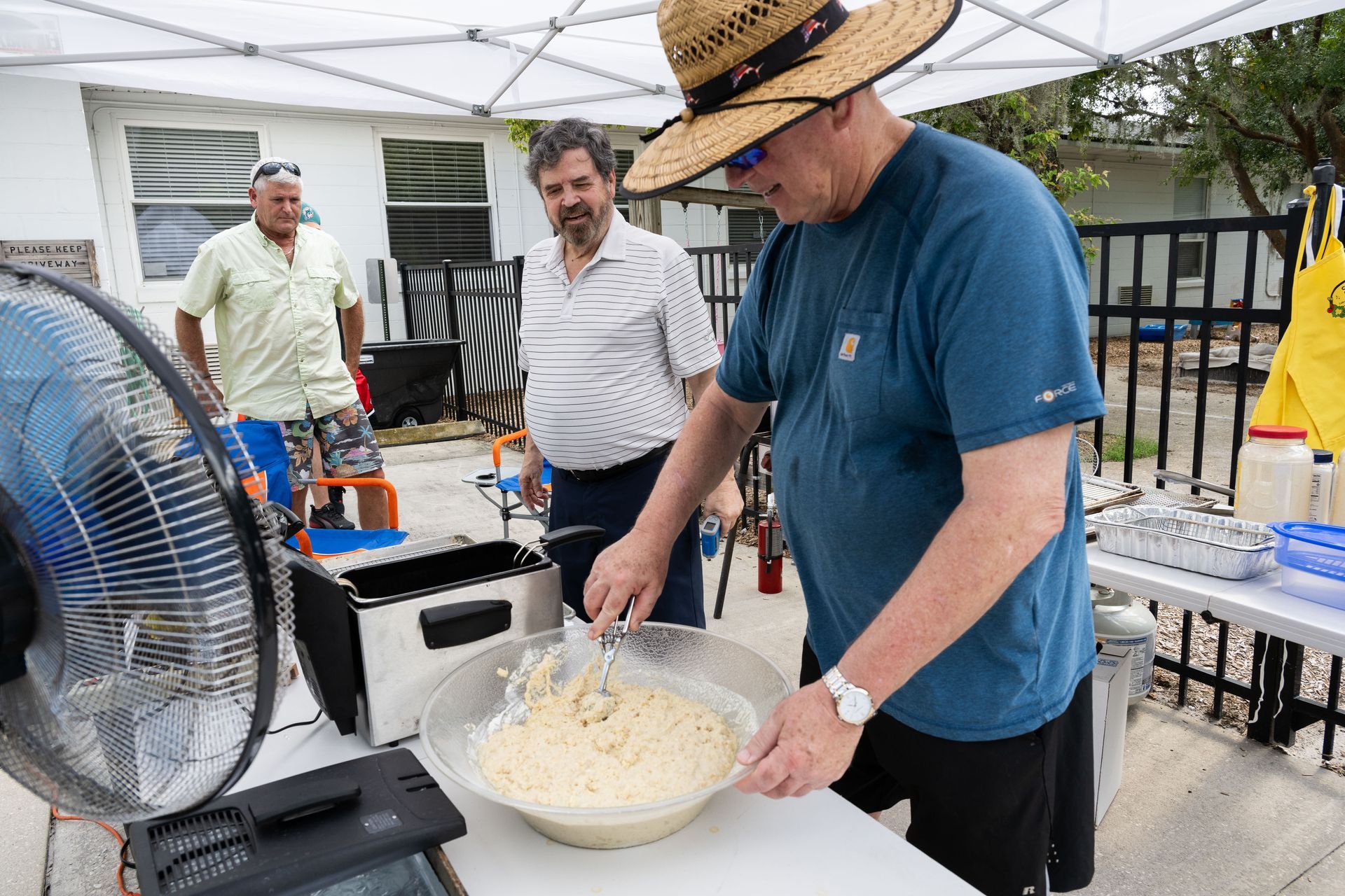 Man stirs batter outside under tent; two men watch. Fan blowing.