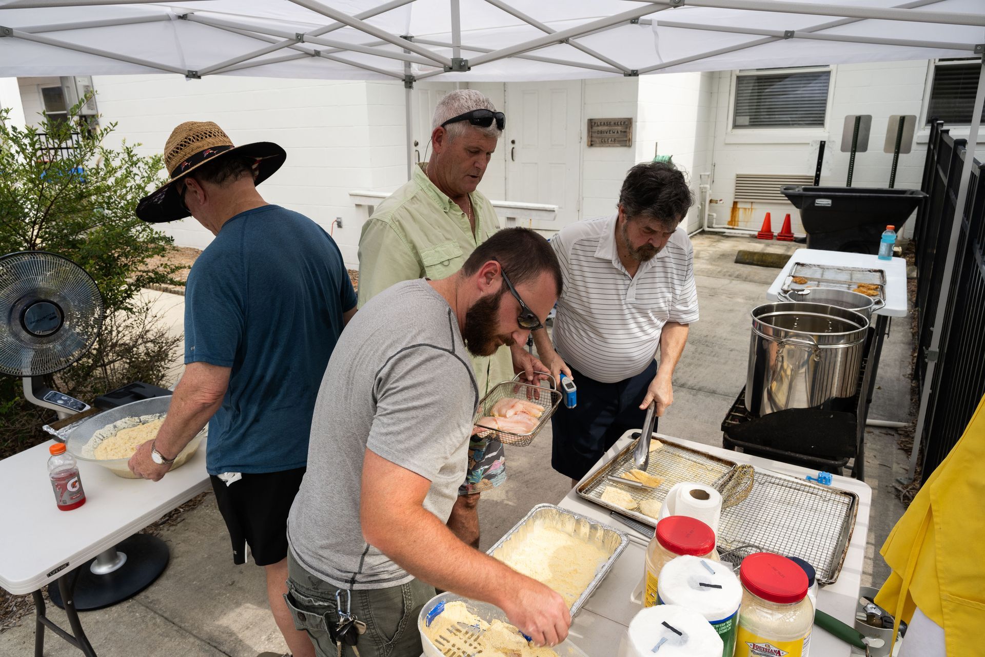 People preparing food at an outdoor event, some near a fryer and tables. A tent provides shade.