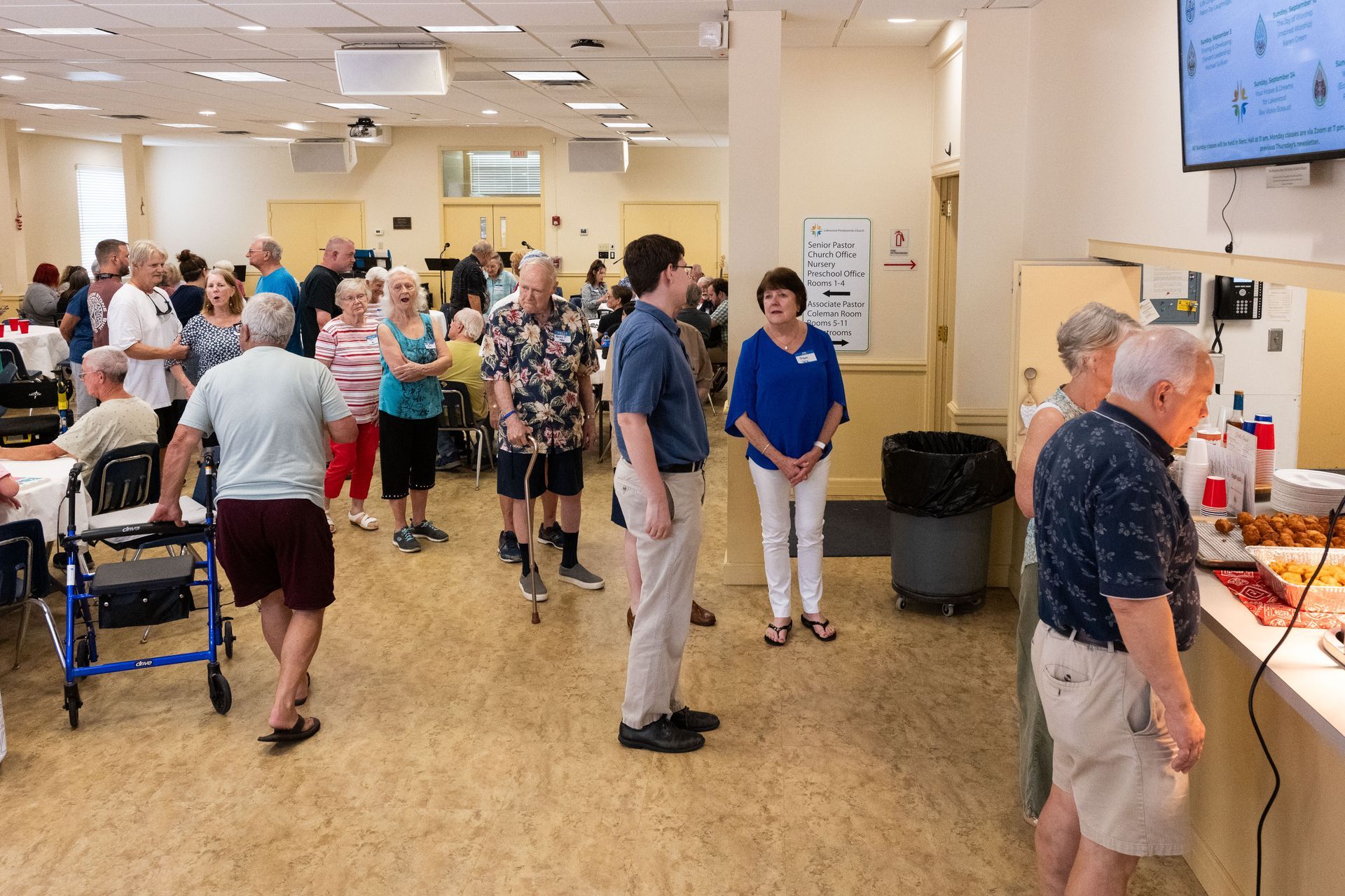 People at an event, some standing in line at a food counter, others socializing. Interior of a brightly lit room.