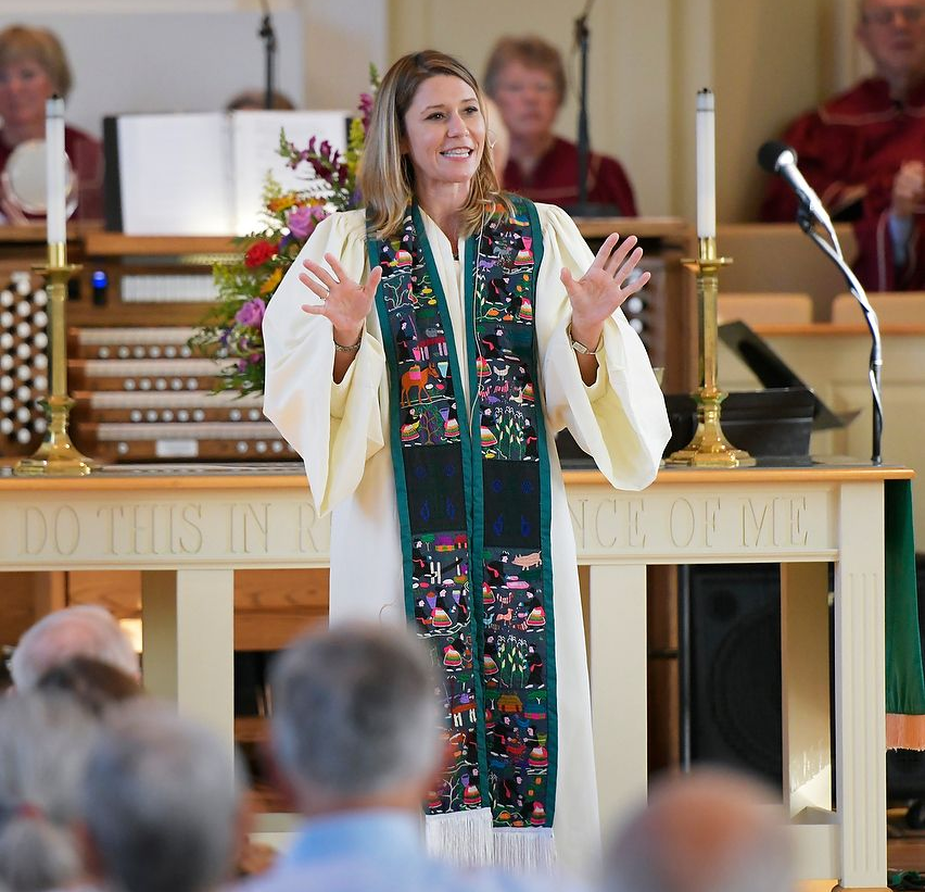Woman in a white robe with a colorful stole, speaking at a church altar, gesturing with hands.