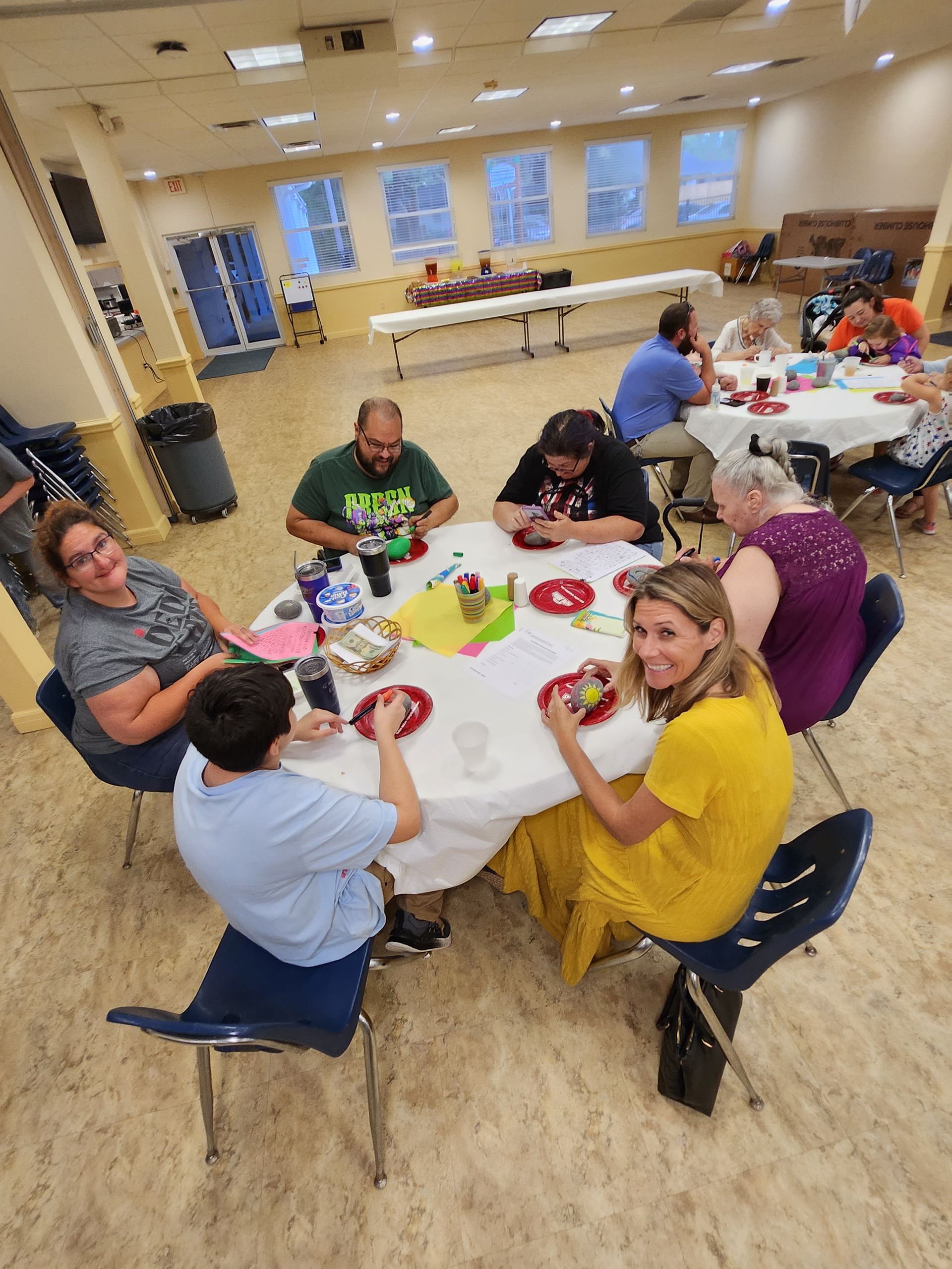 People seated around a table, possibly crafting. The room has a beige floor and white walls.