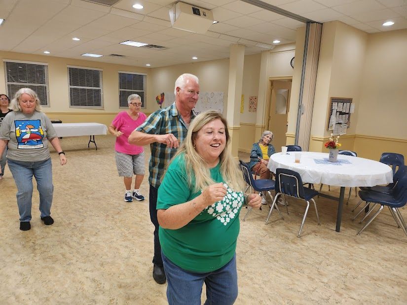 People dancing in a community hall. Woman in green top smiles while dancing with a man. Other dancers follow.