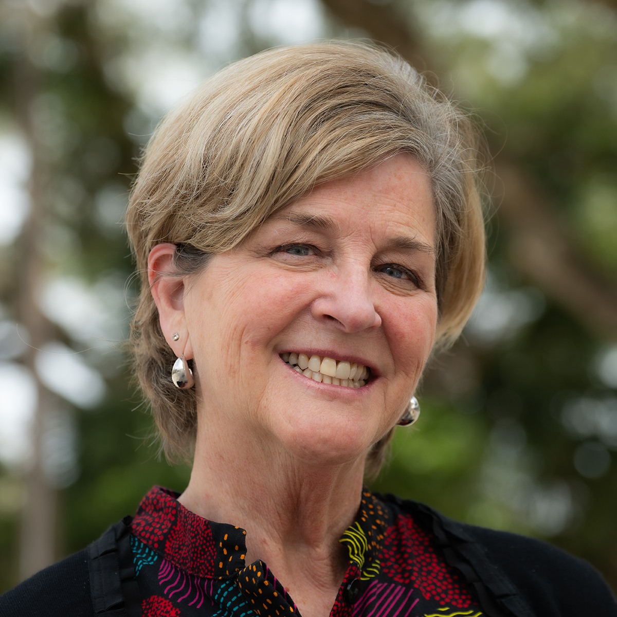 Woman with short blonde hair smiles; she wears a colorful patterned shirt and small silver hoop earrings.