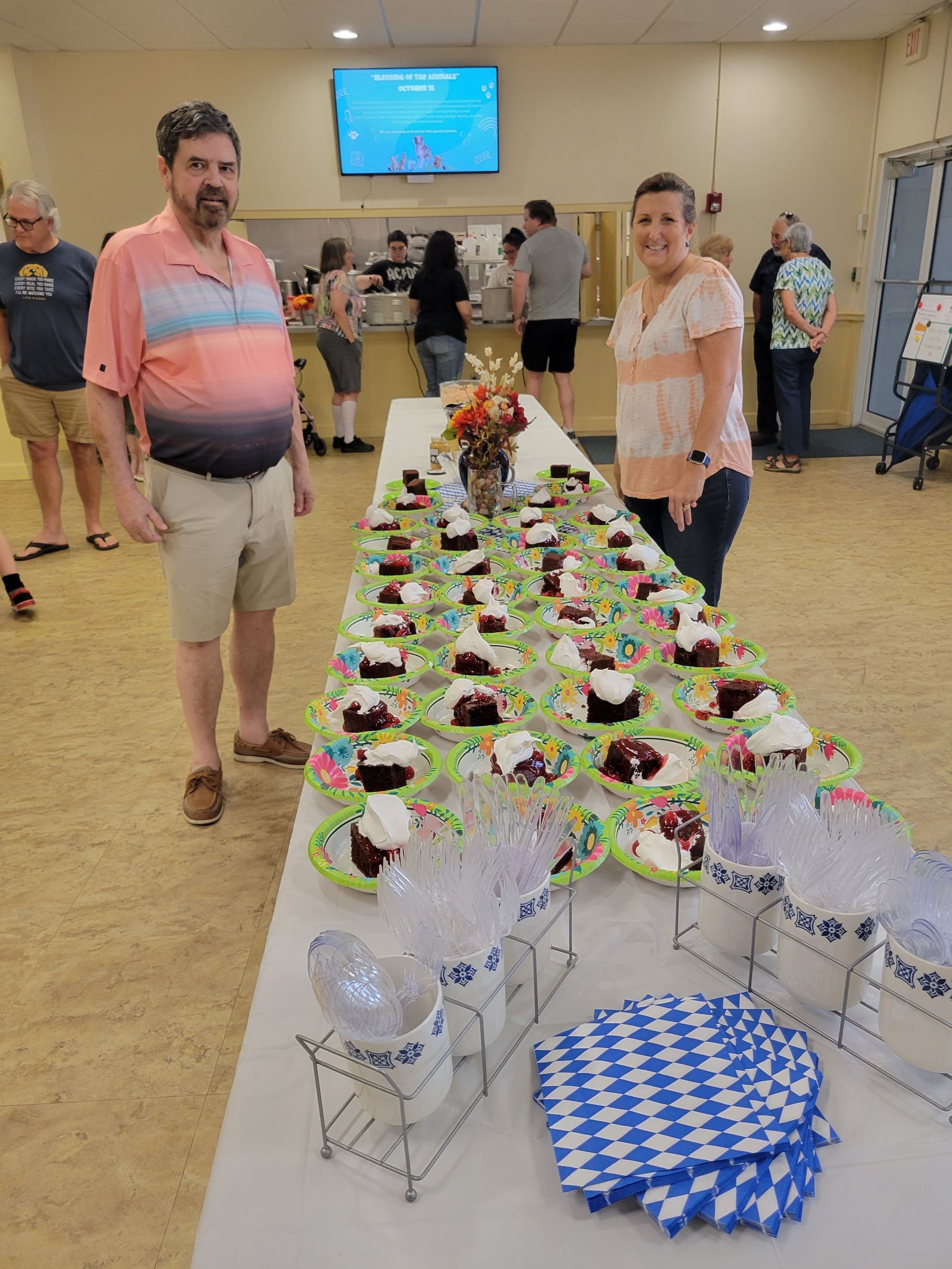 People stand near a long table covered in cupcakes and refreshments at an event in a community center.