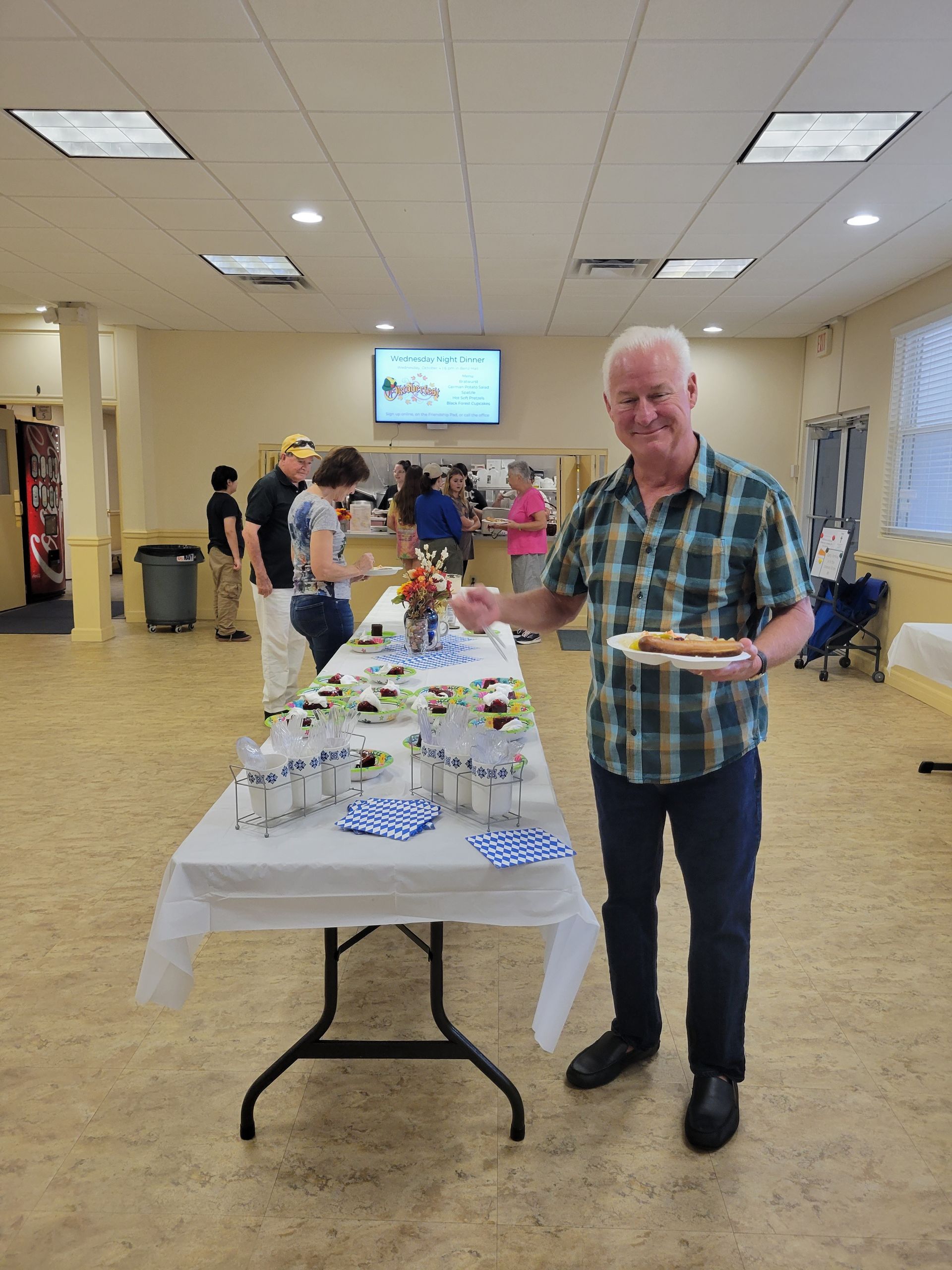 Man with hot dog smiles next to buffet table; others in background, indoor event.