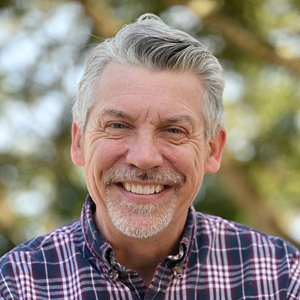 Smiling man with gray hair, wearing a plaid shirt, outdoors with blurred background.