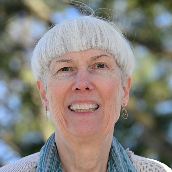 Woman with short, light-colored hair smiles outdoors, wearing a light sweater and scarf; blurred background.