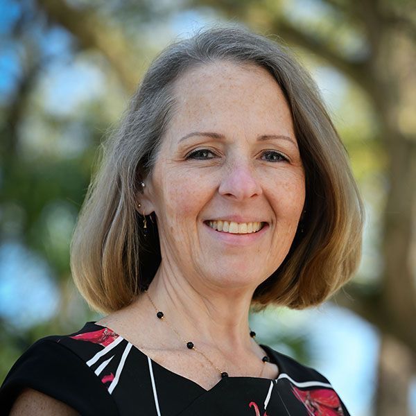 Woman with short gray-blonde hair smiles, wearing a patterned top and black beaded necklace, outdoors.