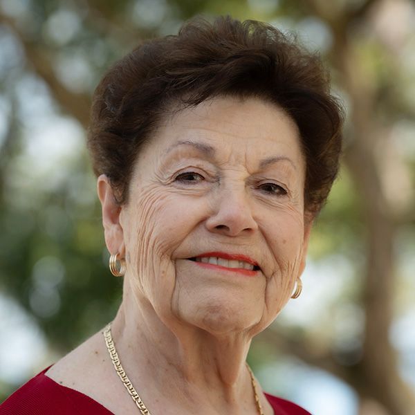 Woman with short brown hair, smiling, wearing gold earrings and necklace; outdoors with a blurred background.