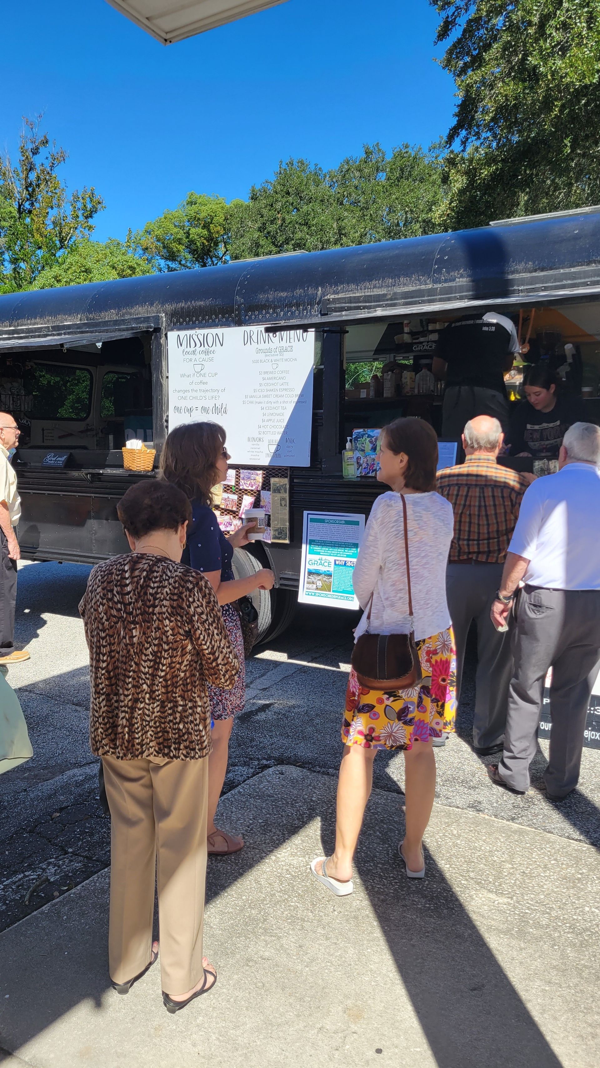 People order food from a black food truck under a bright sky.
