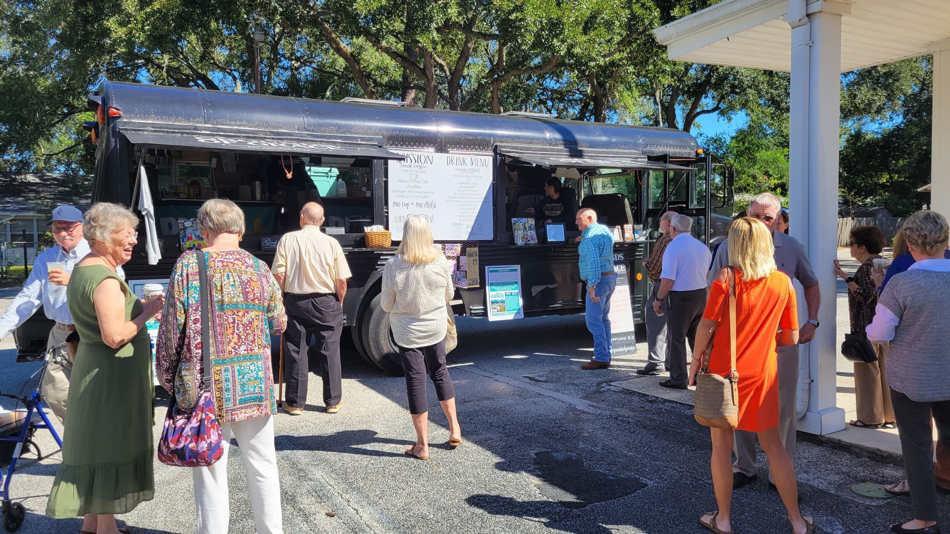 People line up at a dark food truck. Several stand near a white building, some holding drinks.
