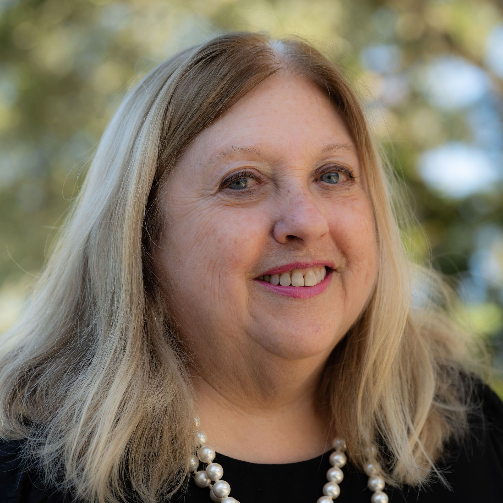 Woman with blonde hair smiles, wearing pearls. Outdoors with blurred background.