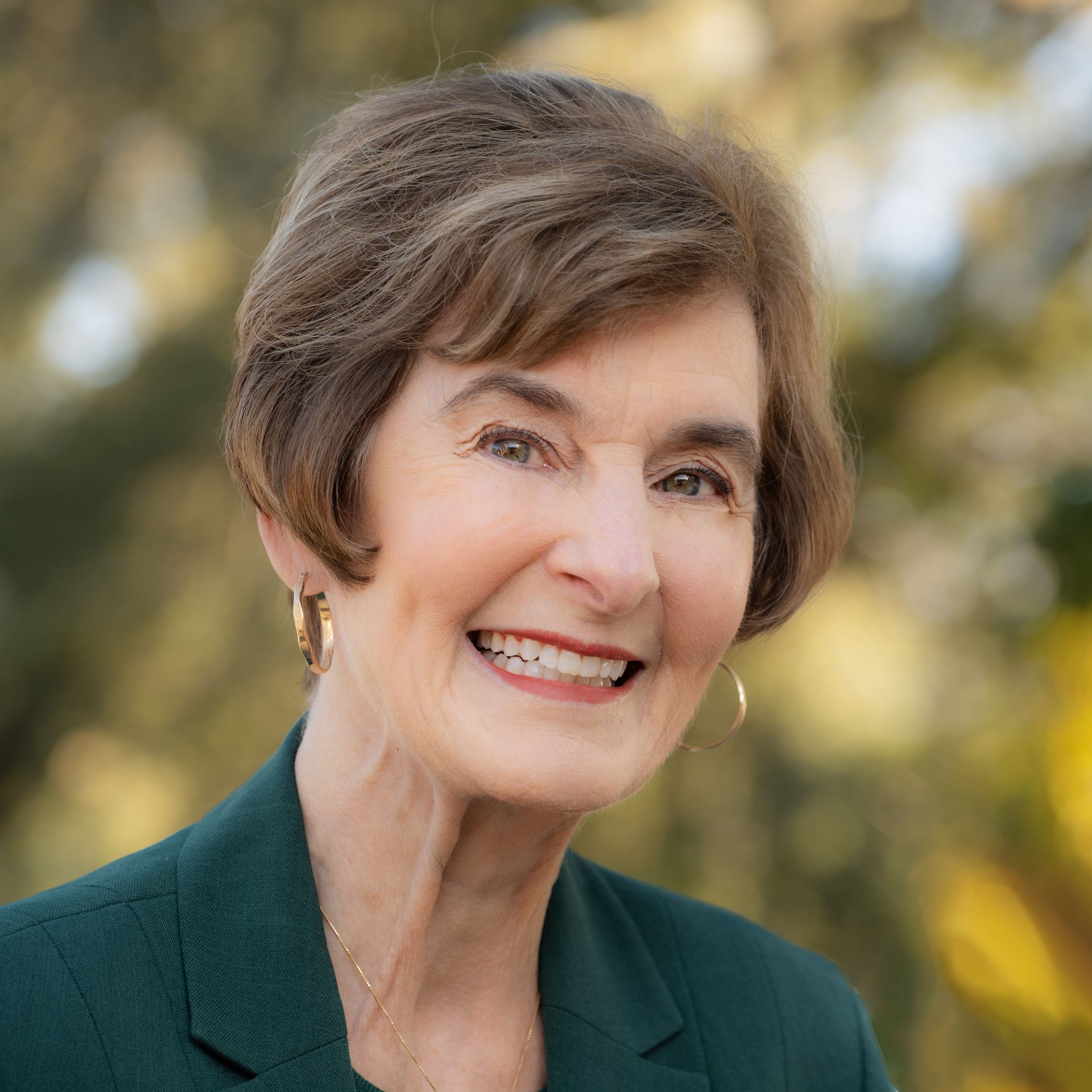 Woman smiling outdoors, wearing a green blazer and gold hoop earrings.