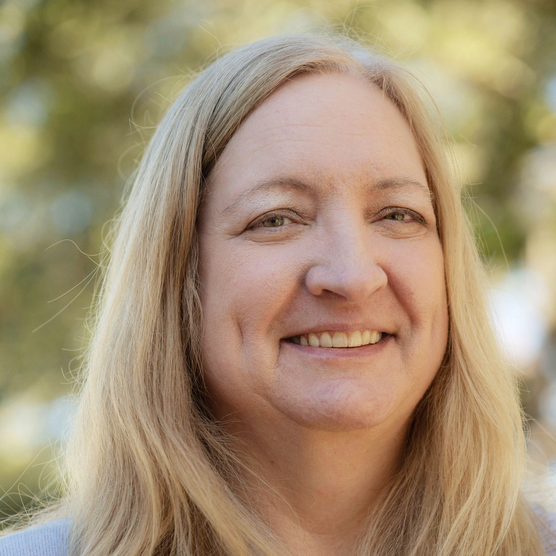 Blonde woman smiling, facing the camera, with a blurred outdoor background.