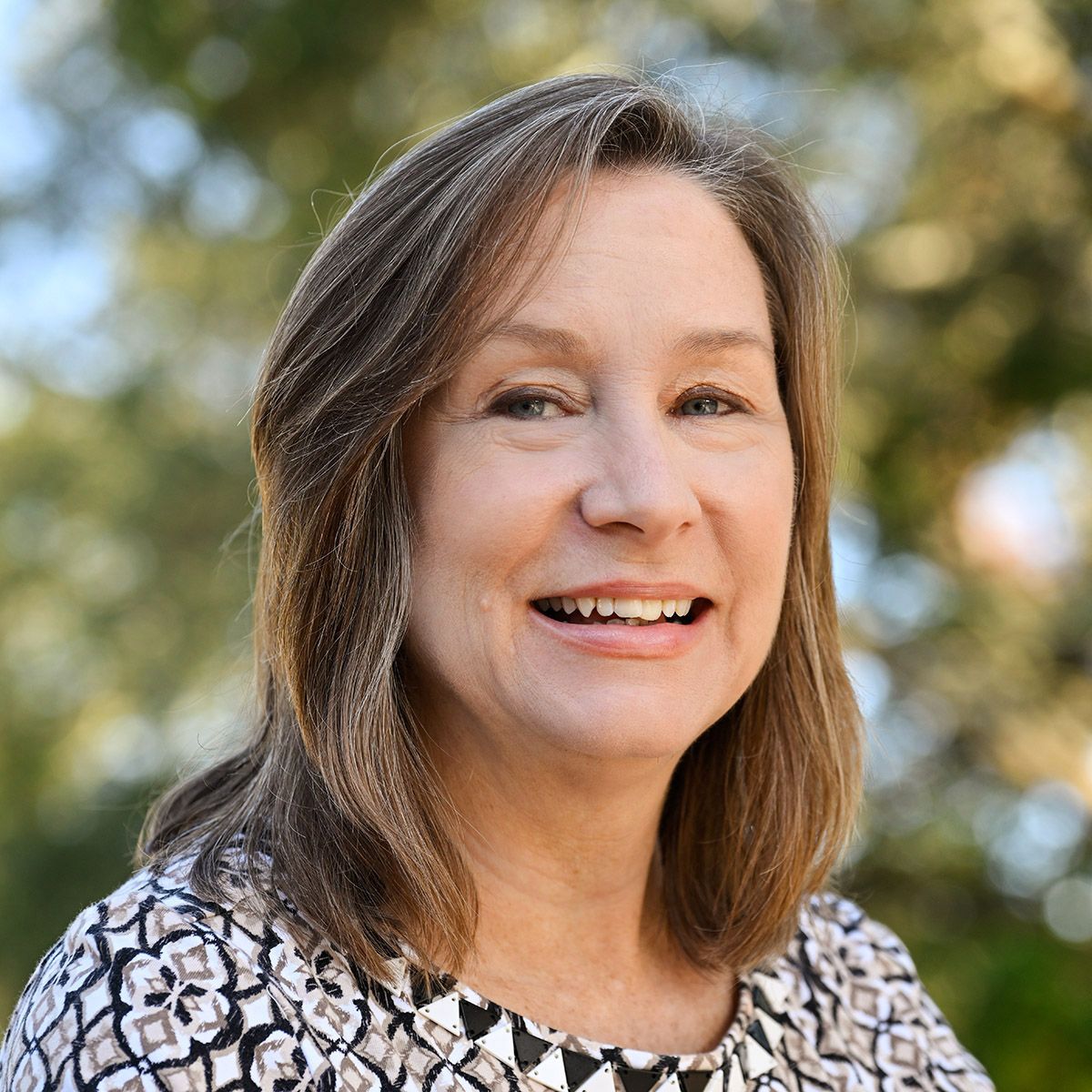 Woman with brown hair smiling, outdoors, neutral background.