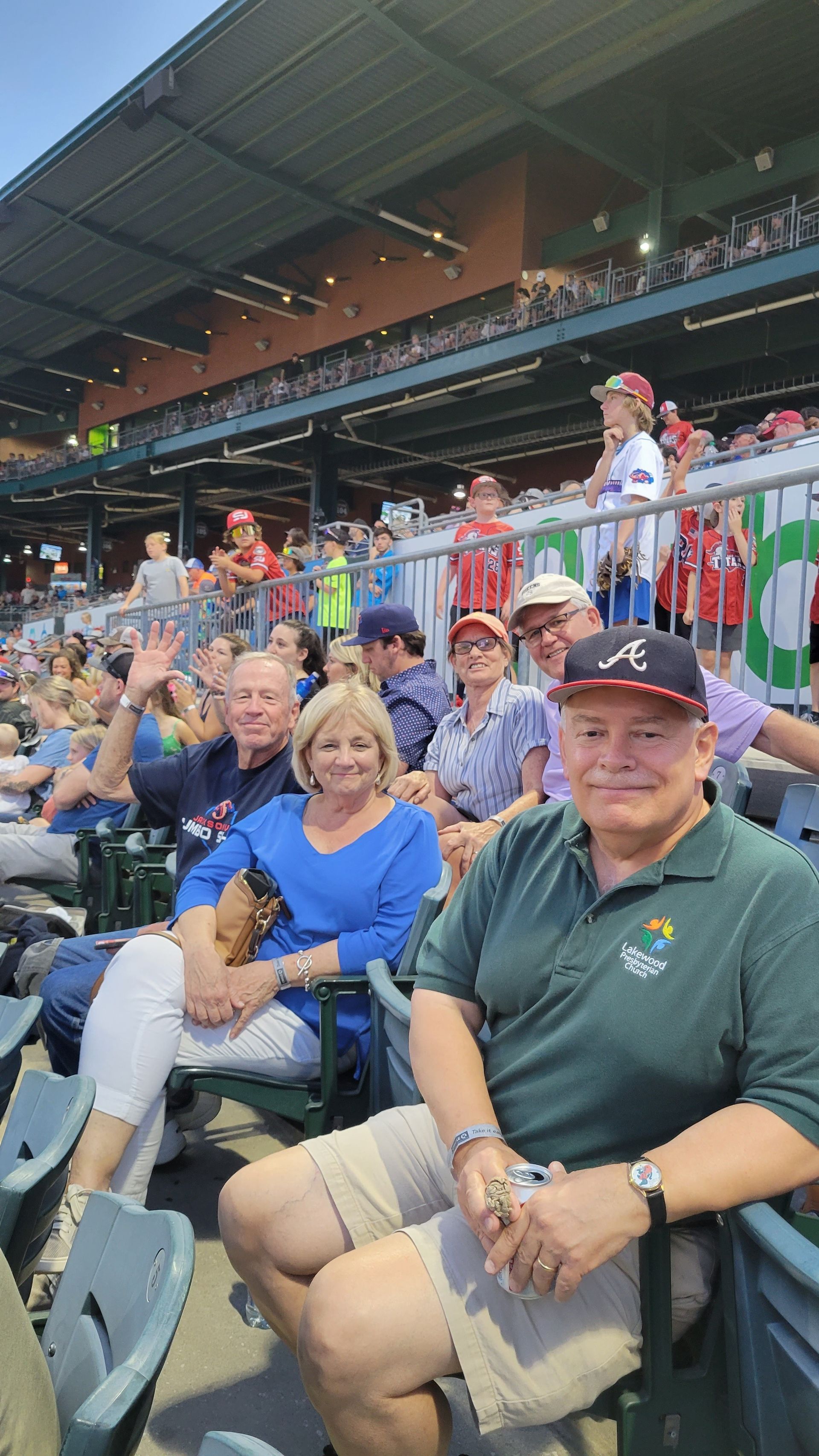 People at a baseball game. Front row: two men and a woman smiling. Others in the seats behind them, also smiling. Stadium in background.