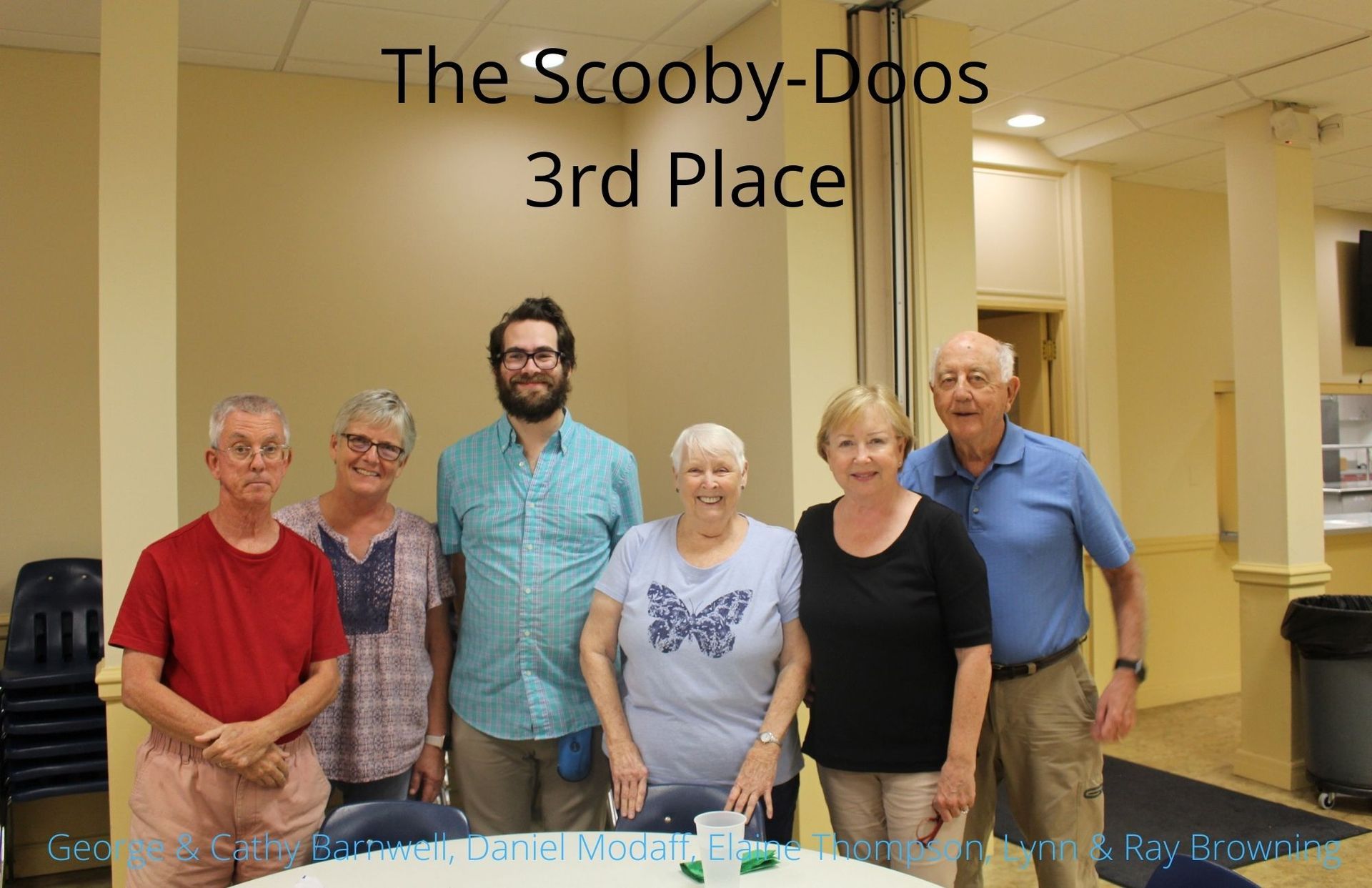 Group of six people standing together, posing for a photo, with a sign that reads 