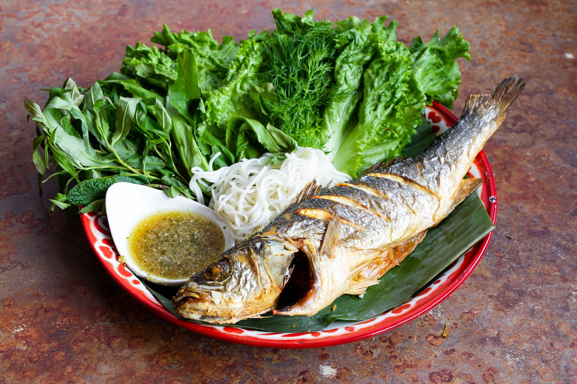 Grilled fish served with greens, vermicelli noodles, and dipping sauce on a plate.