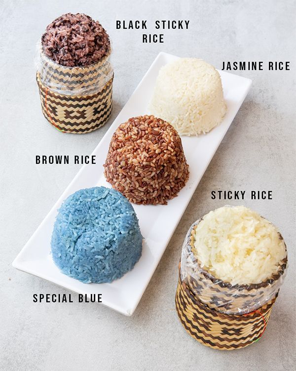 Five types of cooked rice on a white platter: black sticky, jasmine, brown, special blue, and sticky rice, in decorative containers.