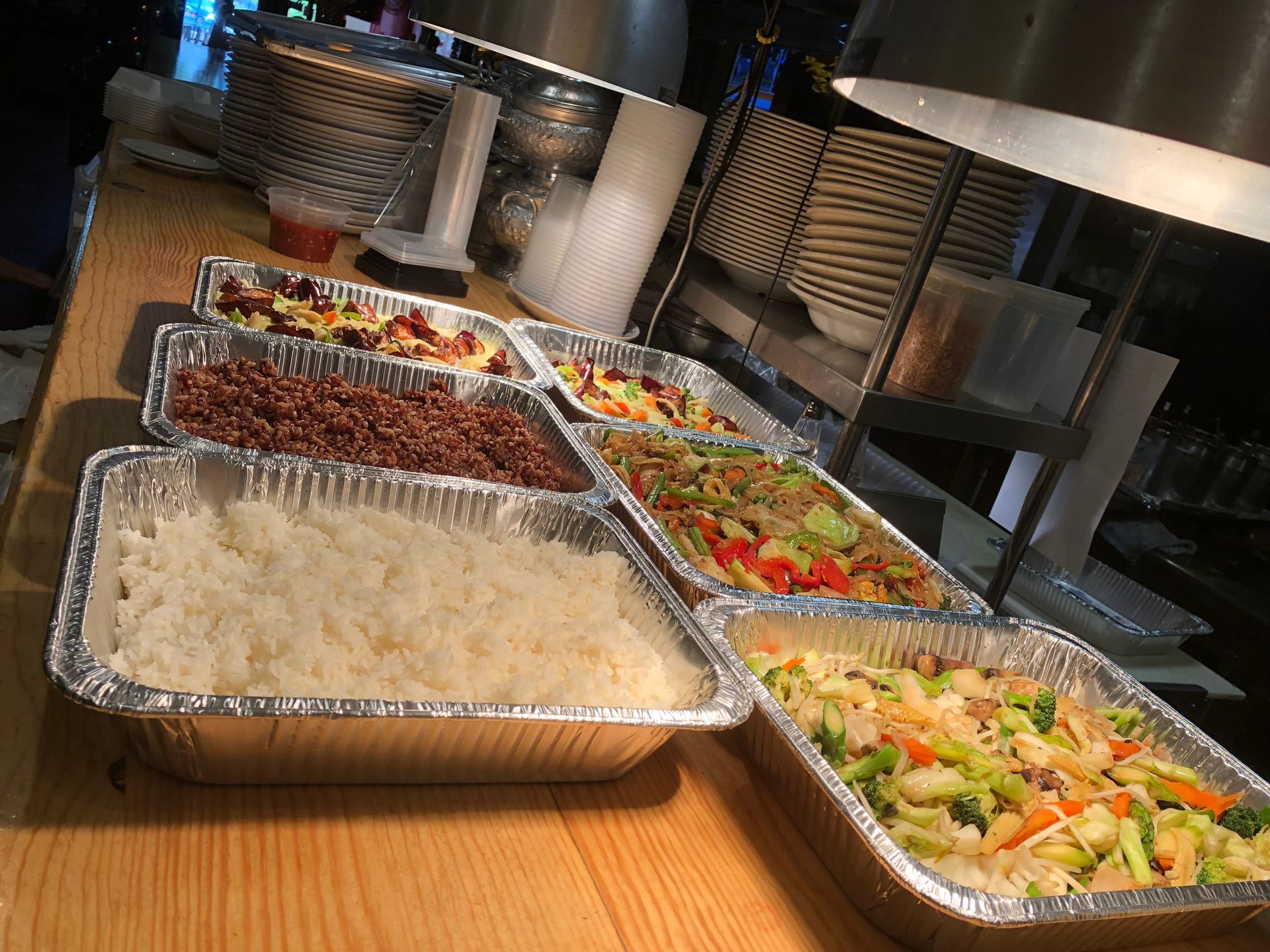 Buffet of various dishes in foil containers, on a wooden counter.