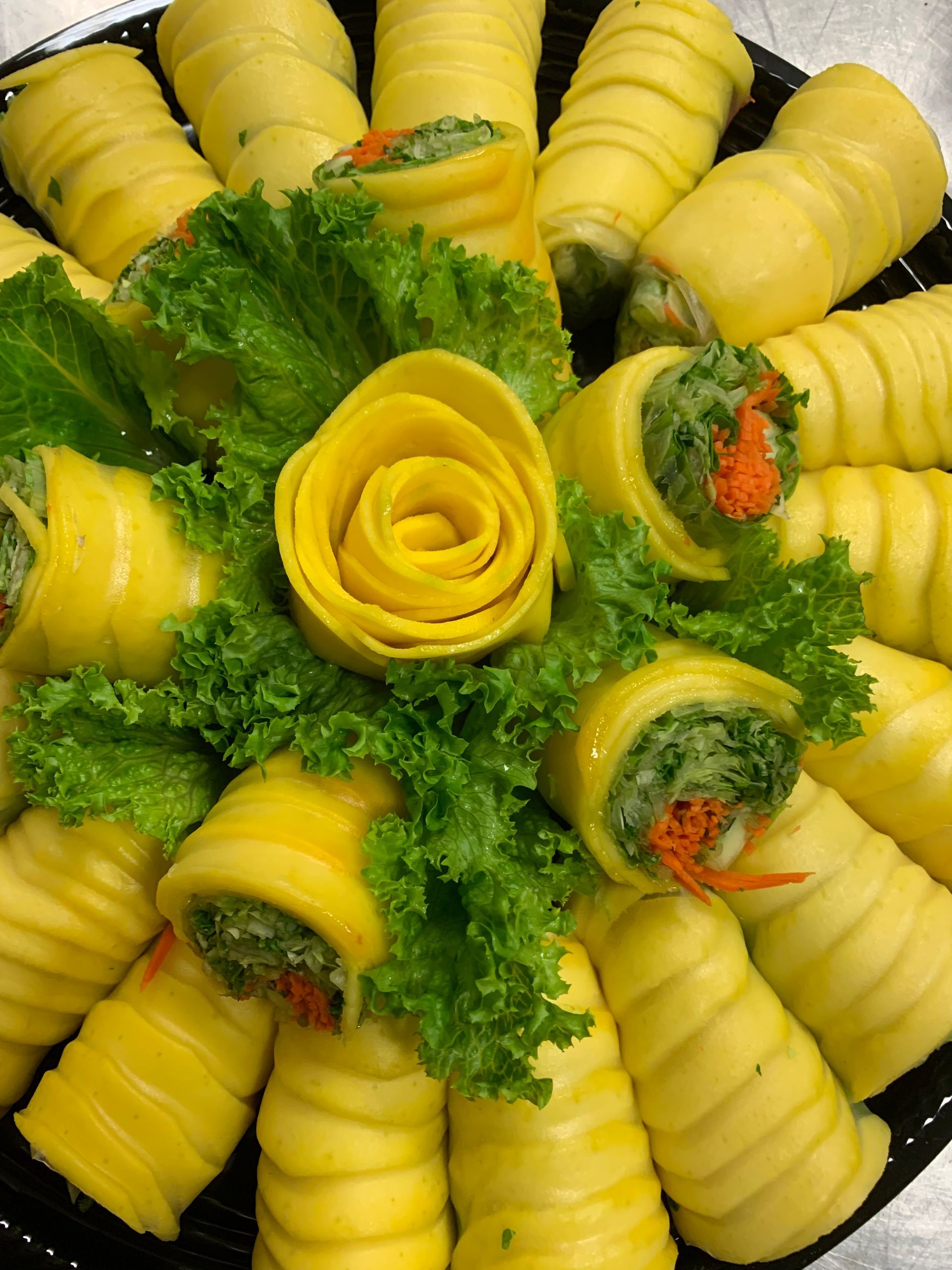 Mango spring rolls with green filling on a platter, surrounded by lettuce and a yellow mango rose garnish.