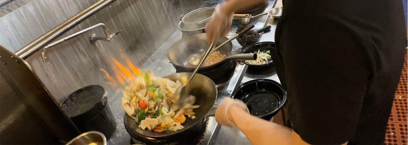 Chef cooking food in a wok with flames visible, likely in a restaurant kitchen.