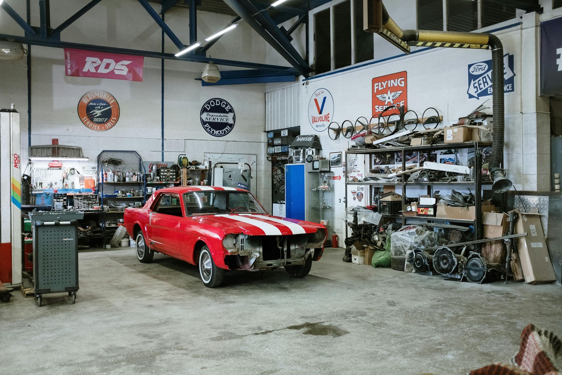 Red car in a garage with white racing stripes. Tools and parts on shelves, banners on walls.
