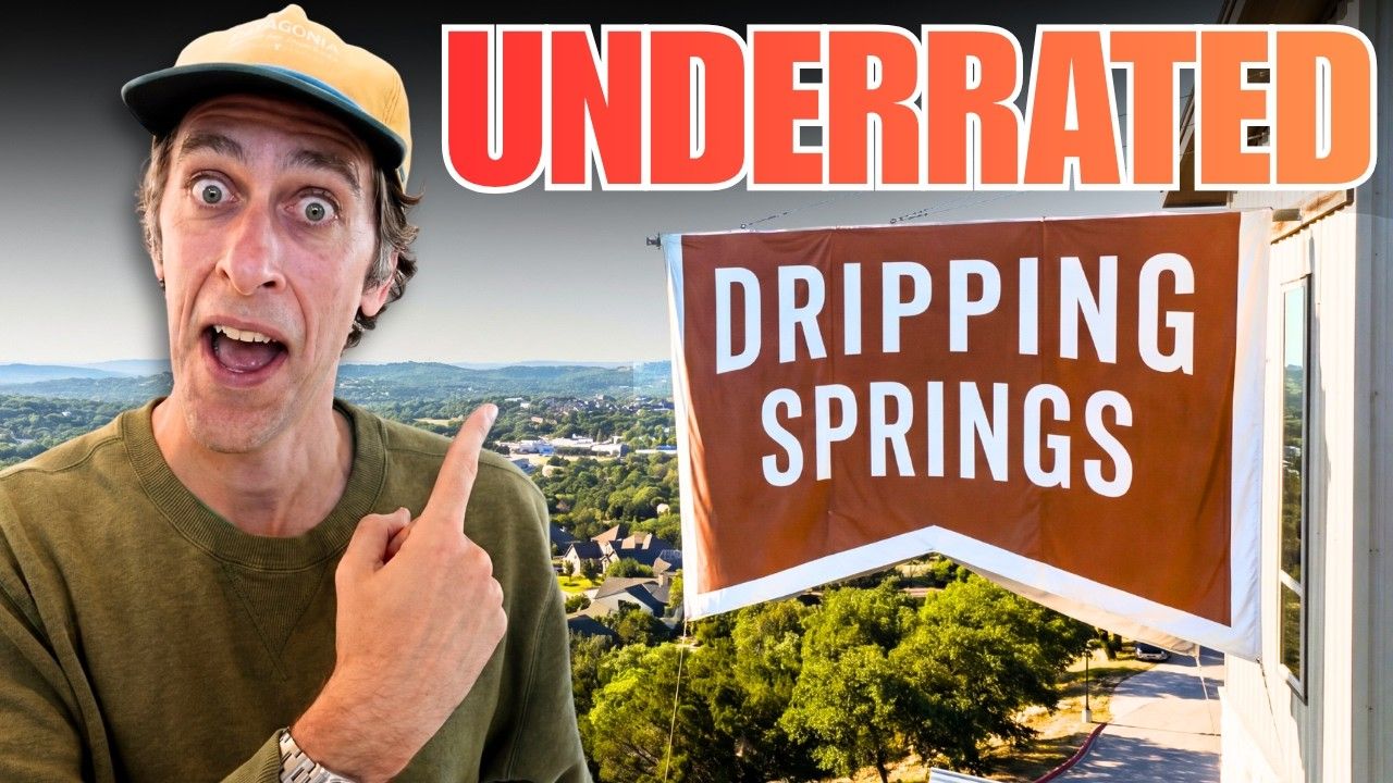 Man pointing at “Underrated Dripping Springs” sign with scenic town view behind him