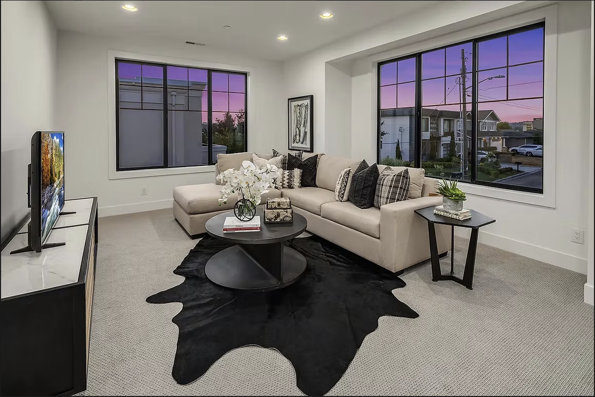 Modern living room with neutral colors, large windows with sunset view, dark rug, and stylish furniture.