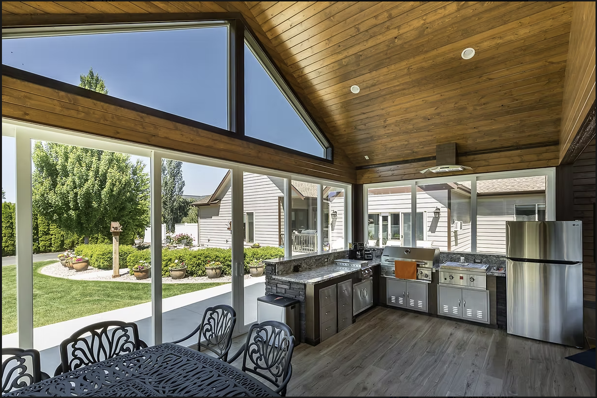Outdoor kitchen with stainless steel appliances, stone countertop, and wooden ceiling, overlooking a backyard.