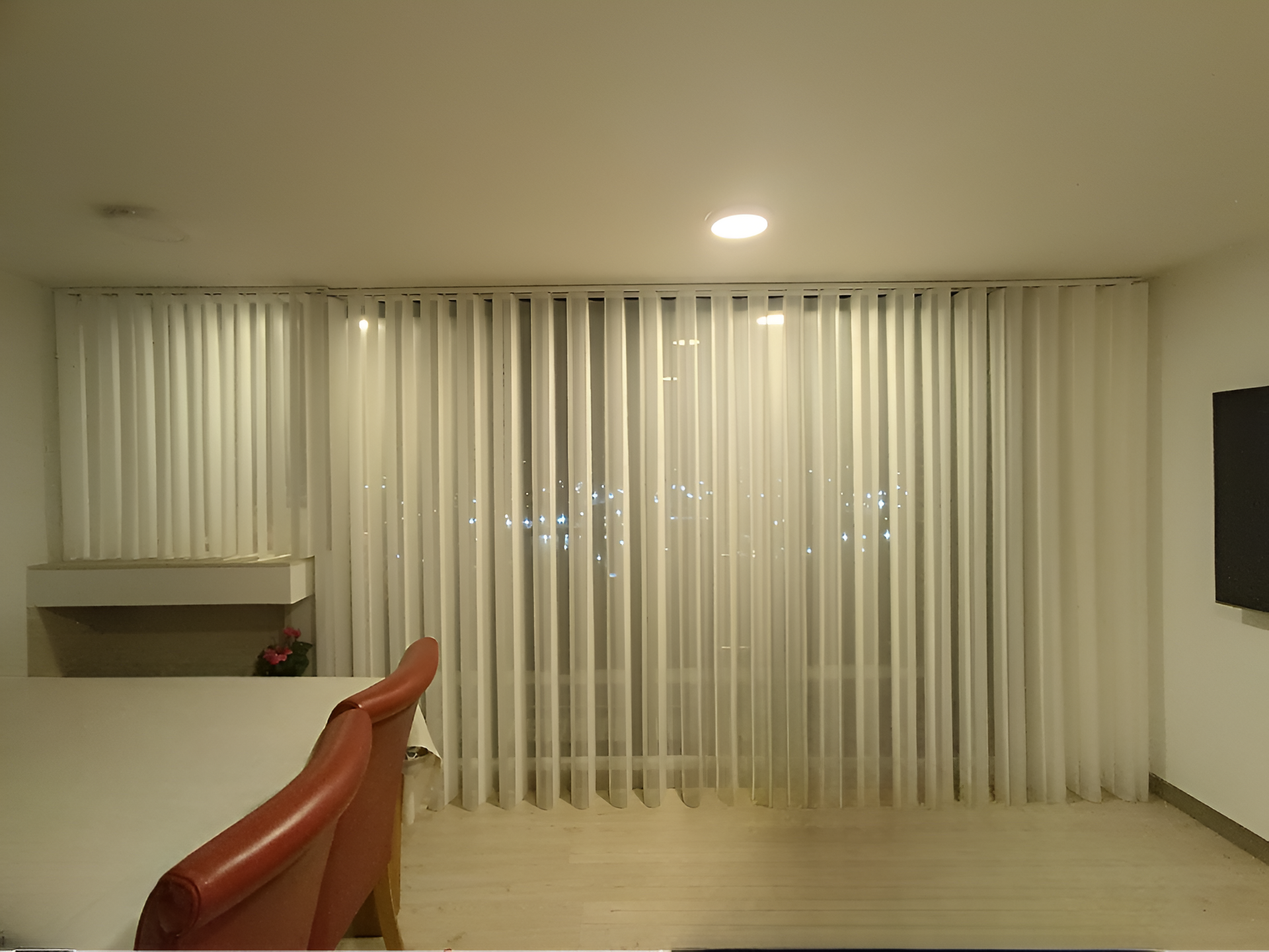 Cortinas plisadas de color beige que cuelgan de una barra de metal oscuro, con una pared gris al fondo.