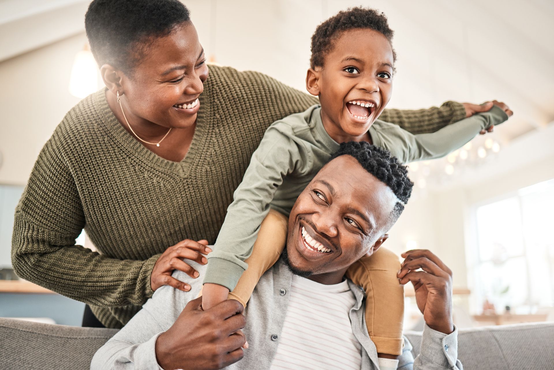 Family, smiling, having fun. Boy on father's shoulders, mother smiling next to them, in a living room.