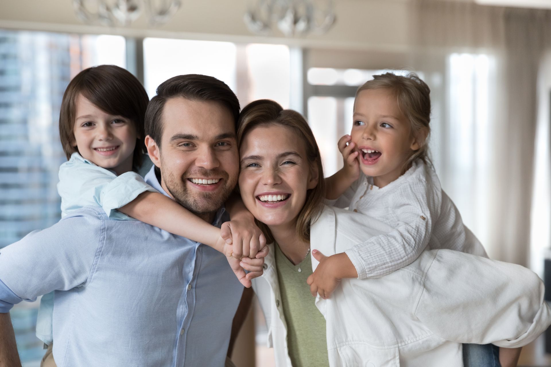 Family of four smiling, posing indoors. Children on parents' backs. Natural light.
