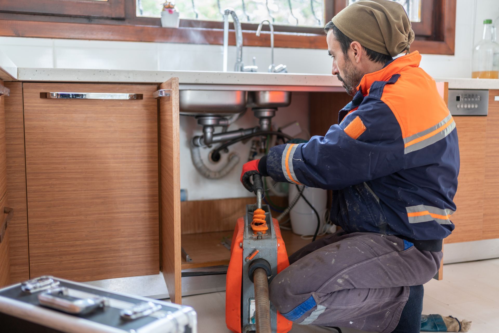 A plumber in uniform fixing a clogged kitchen sink pipe under the counter with tools. A plumber in uniform fixing a clogged kitchen sink pipe under the counter with tools.