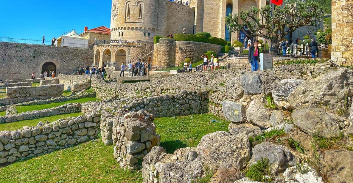 A historic site with stone ruins and fortress walls under a bright blue sky. Tourists walk through the area, exploring the ancient stone structures. The site is surrounded by greenery and overlooks a well-preserved castle or fort in the background.