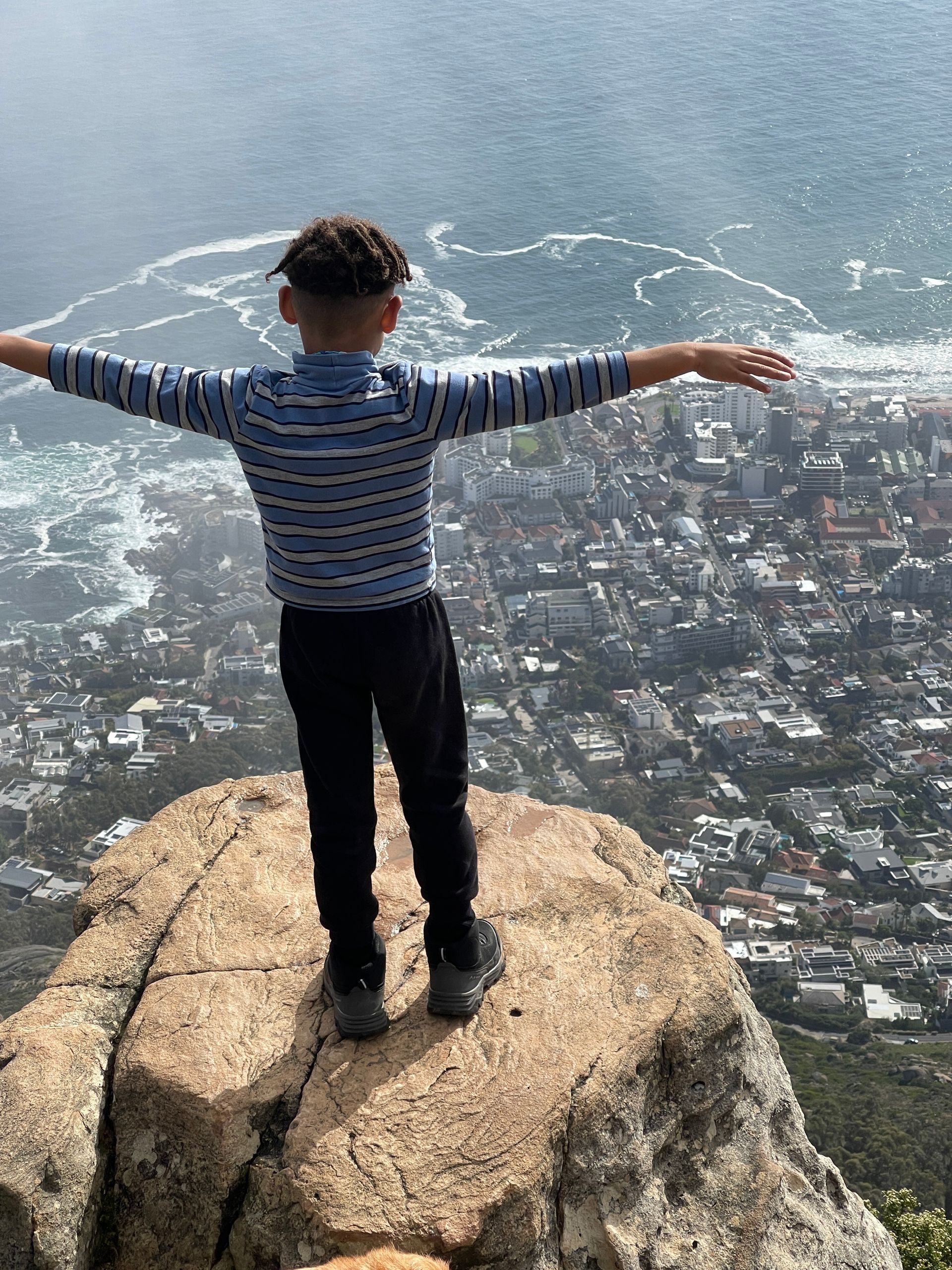 A young boy is standing on top of a rock with his arms outstretched o Lions Head Mountain in Cape T