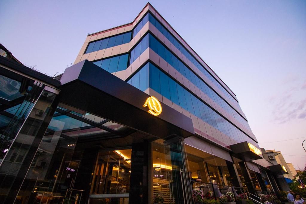 A modern glass-fronted building with reflective windows and sleek architectural design. The entrance is highlighted by a black awning with a gold logo, with the evening sky casting a soft light over the structure.