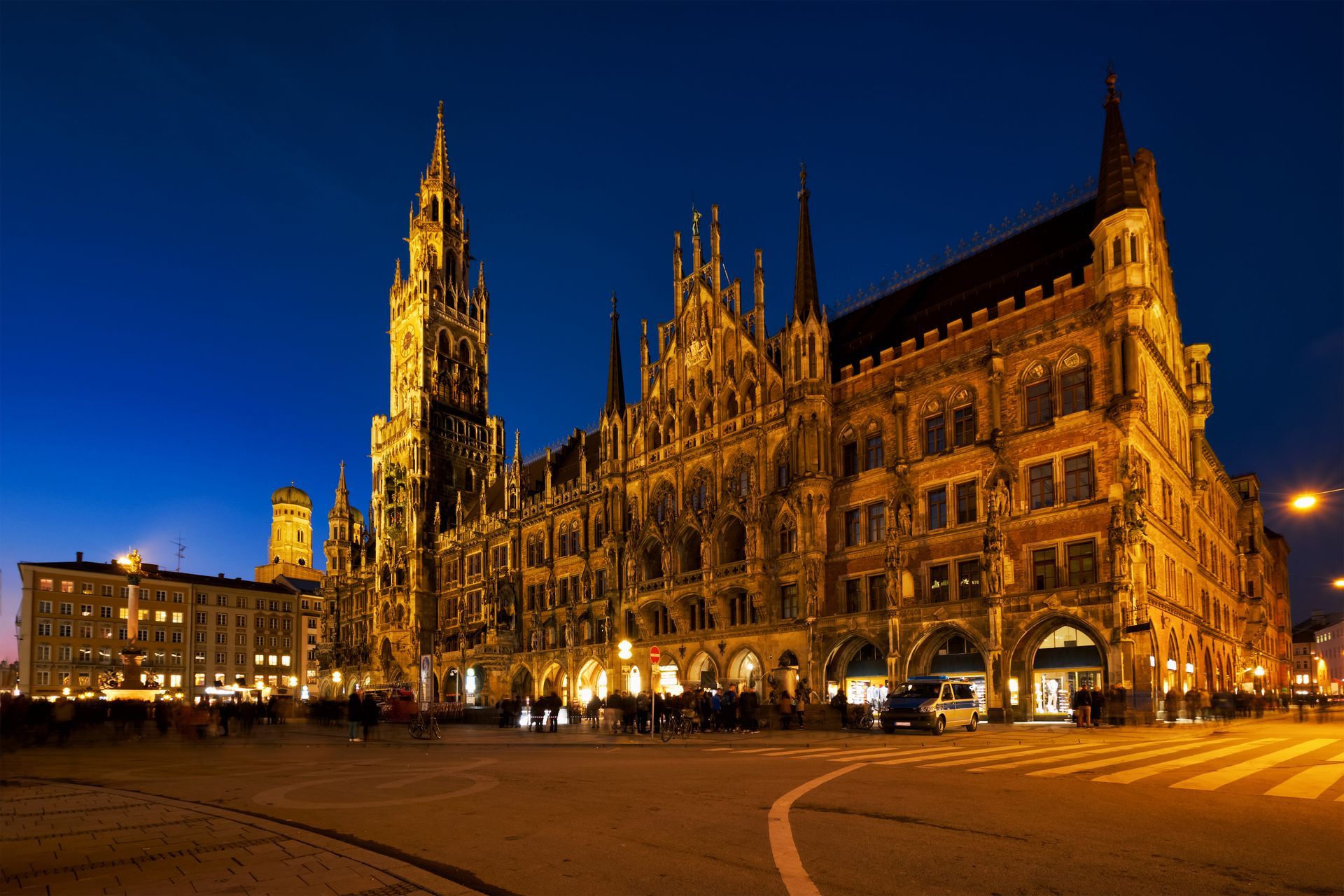 The image shows the New Town Hall (Neues Rathaus) in Munich, Germany, illuminated at night. The neo-gothic building is located in Marienplatz, the city's central square, and is notable for its intricate façade with pointed towers and spires. The tower houses the famous Glockenspiel, a mechanical clock that features life-sized figures. The sky is a deep blue, and the warm lighting of the building creates a striking contrast against the evening backdrop. There are people and cars in the square, adding to the vibrant urban atmosphere.