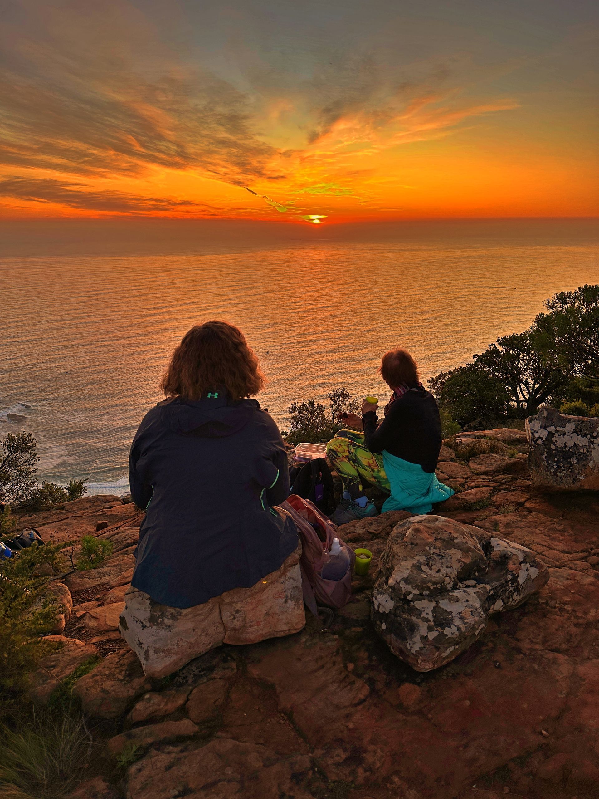 Two people sitting on a rocky cliff, watching a beautiful sunset over the ocean. The sky is painted in shades of orange, pink, and purple, and the sun is just above the horizon, creating a peaceful and serene moment as they enjoy the view.