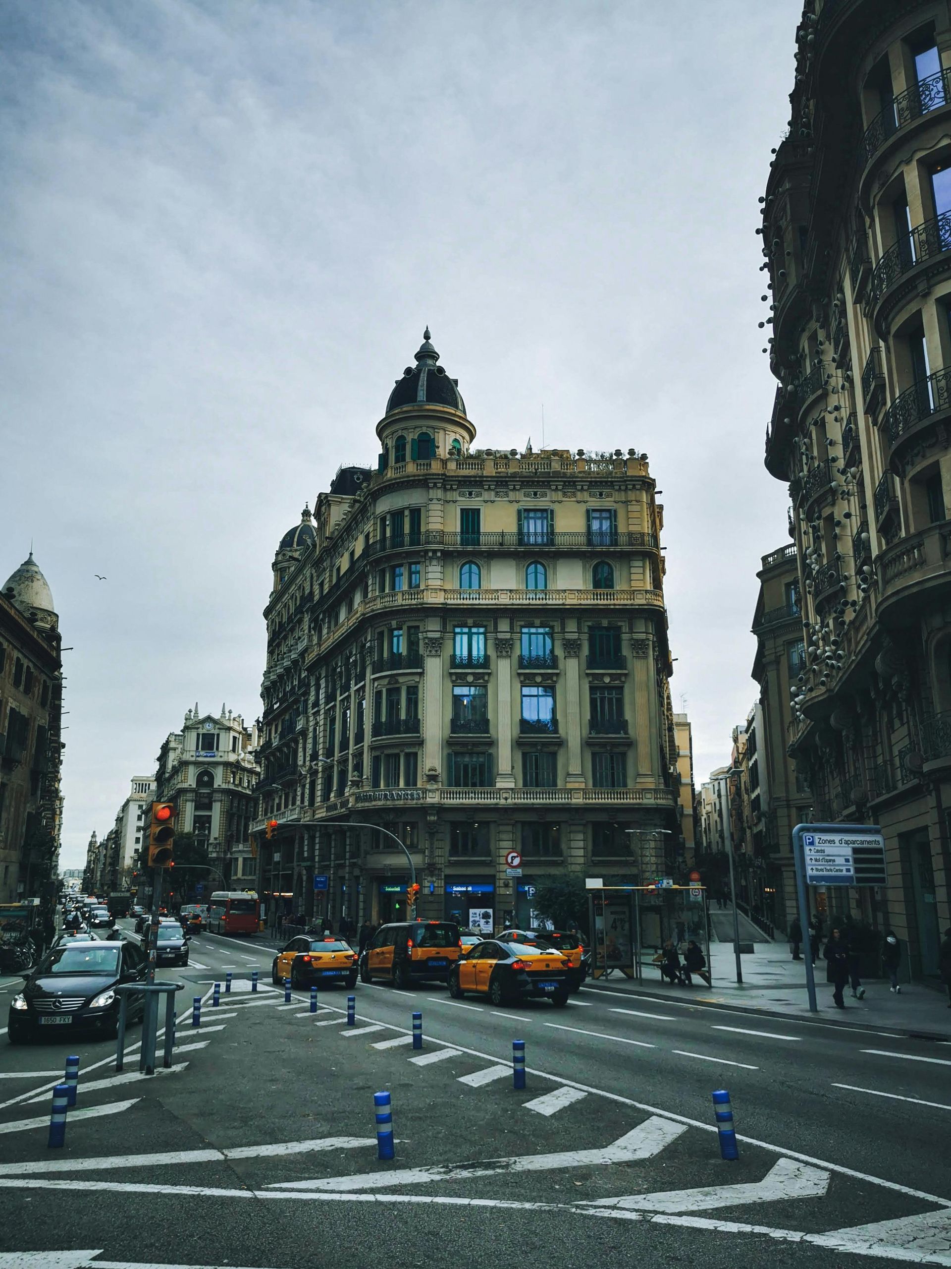 A bustling city street in Barcelona, Spain, lined with grand historic buildings. Yellow taxis and cars are seen at the intersection, with pedestrians walking along the sidewalks. The overcast sky adds a moody atmosphere to the urban scene Barcelona