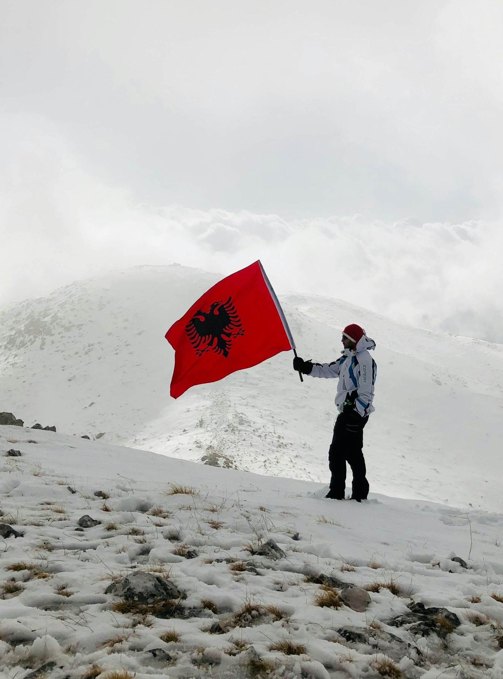 A hiker standing on a snowy mountain slope, holding an Albanian flag featuring a black double-headed eagle on a red background. The surrounding landscape is misty, with snow-covered peaks in the distance under a cloudy sky.