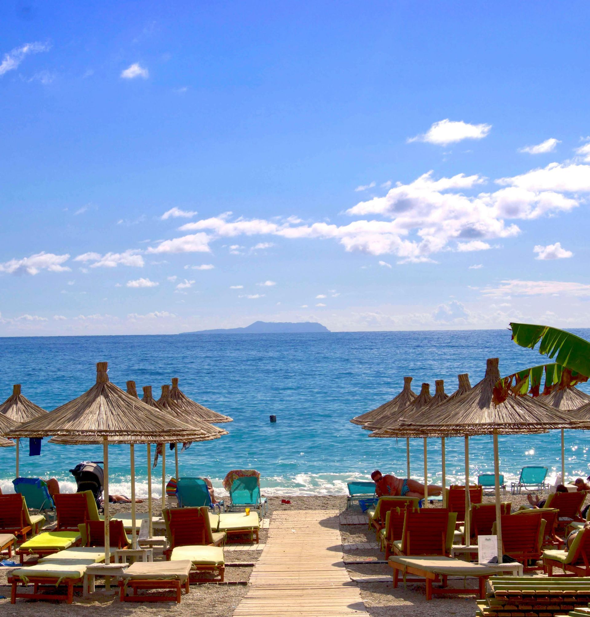 A sunny beach with wooden sun loungers and straw umbrellas arranged along the shore. A wooden path leads to the calm blue ocean, with an island visible on the horizon under a clear, blue sky.