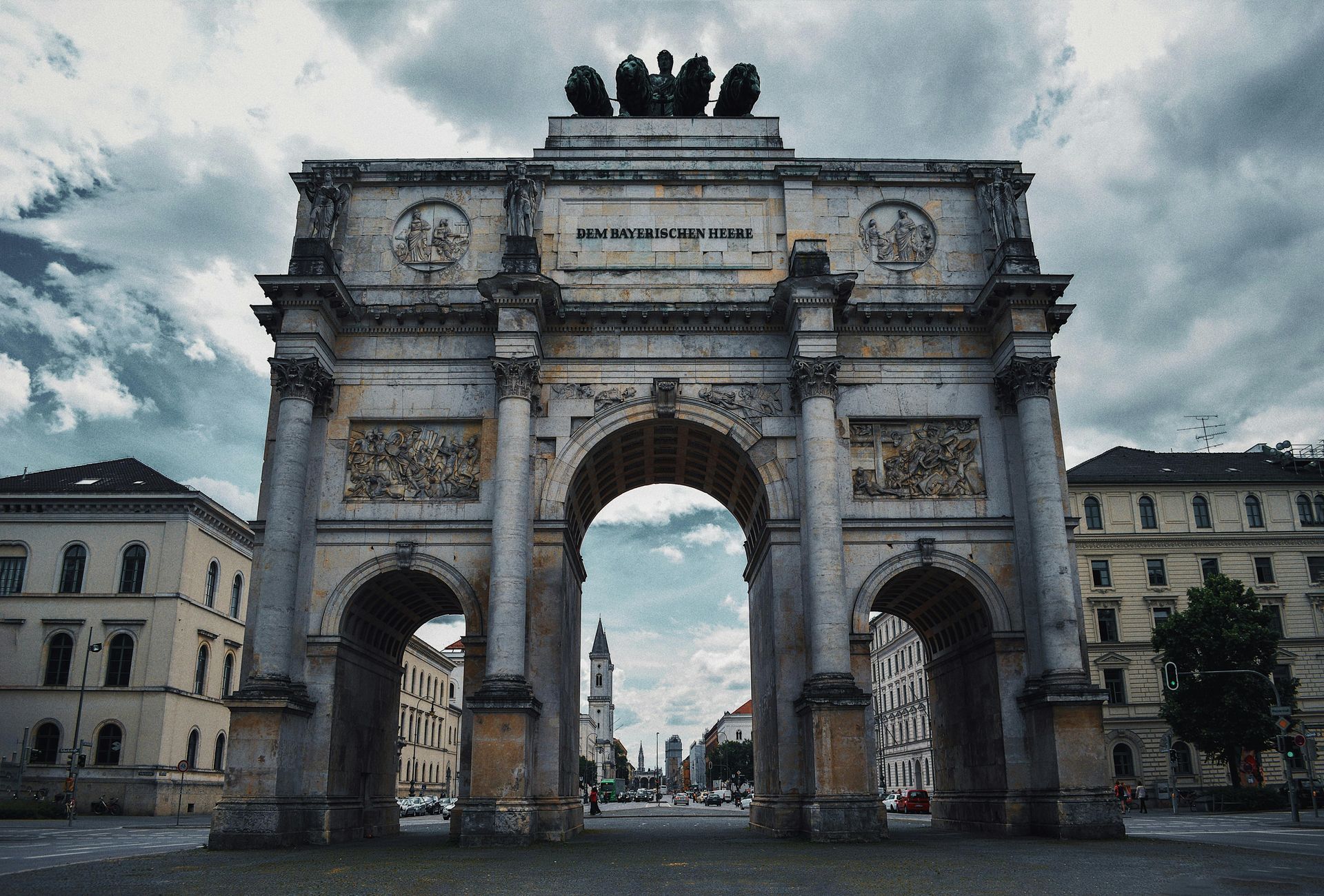 The image shows the Siegestor (Victory Gate) in Munich, Germany. It is a triumphal arch located on Leopoldstraße, originally built to celebrate the Bavarian army's victory. The structure has three large arches, and a statue of four lions pulling a chariot can be seen at the top. The architectural style is neoclassical, and the scene features a cloudy sky and the surrounding buildings typical of the city's urban setting.