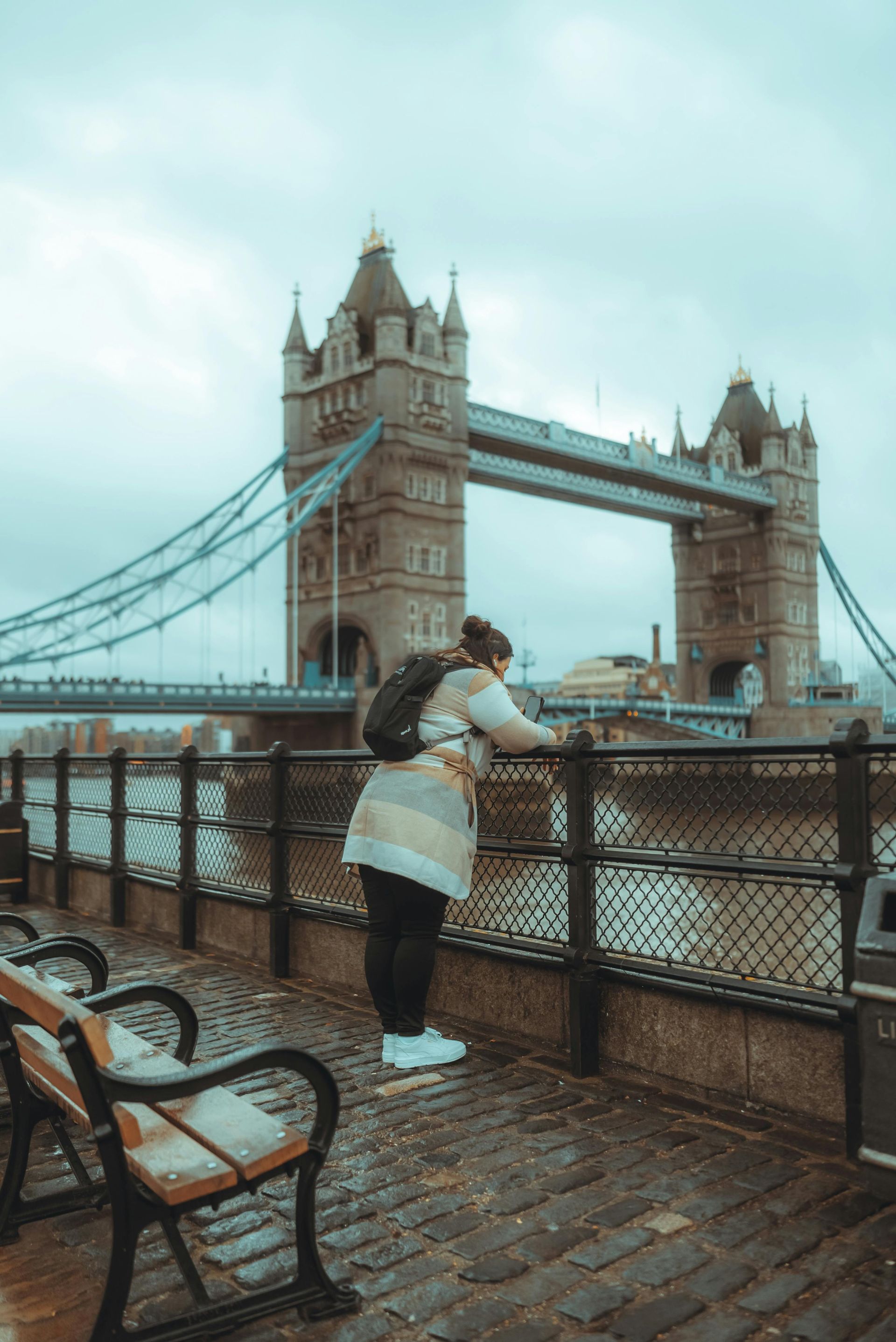 A person wearing a backpack, standing on a cobblestone path by the river, gazing at London's Tower Bridge. The scene is misty with overcast skies, and the iconic bridge stands tall in the background, surrounded by water and a metal railing