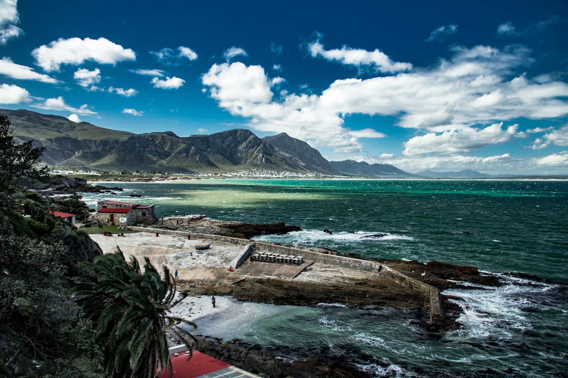 A dramatic coastal view showing rugged shorelines against a backdrop of mountains. The ocean is choppy, and the sky is partly cloudy, enhancing the dramatic effect of the natural scenery. Small buildings near the shore add a human element to the wild landscape.