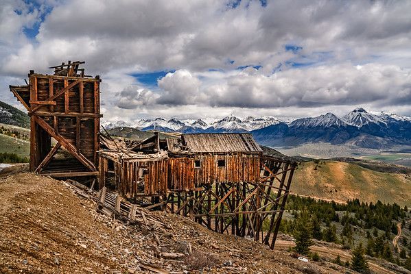 Dilapidated wooden structure on a hillside, possibly an old mine, with mountains and cloudy sky in the background.